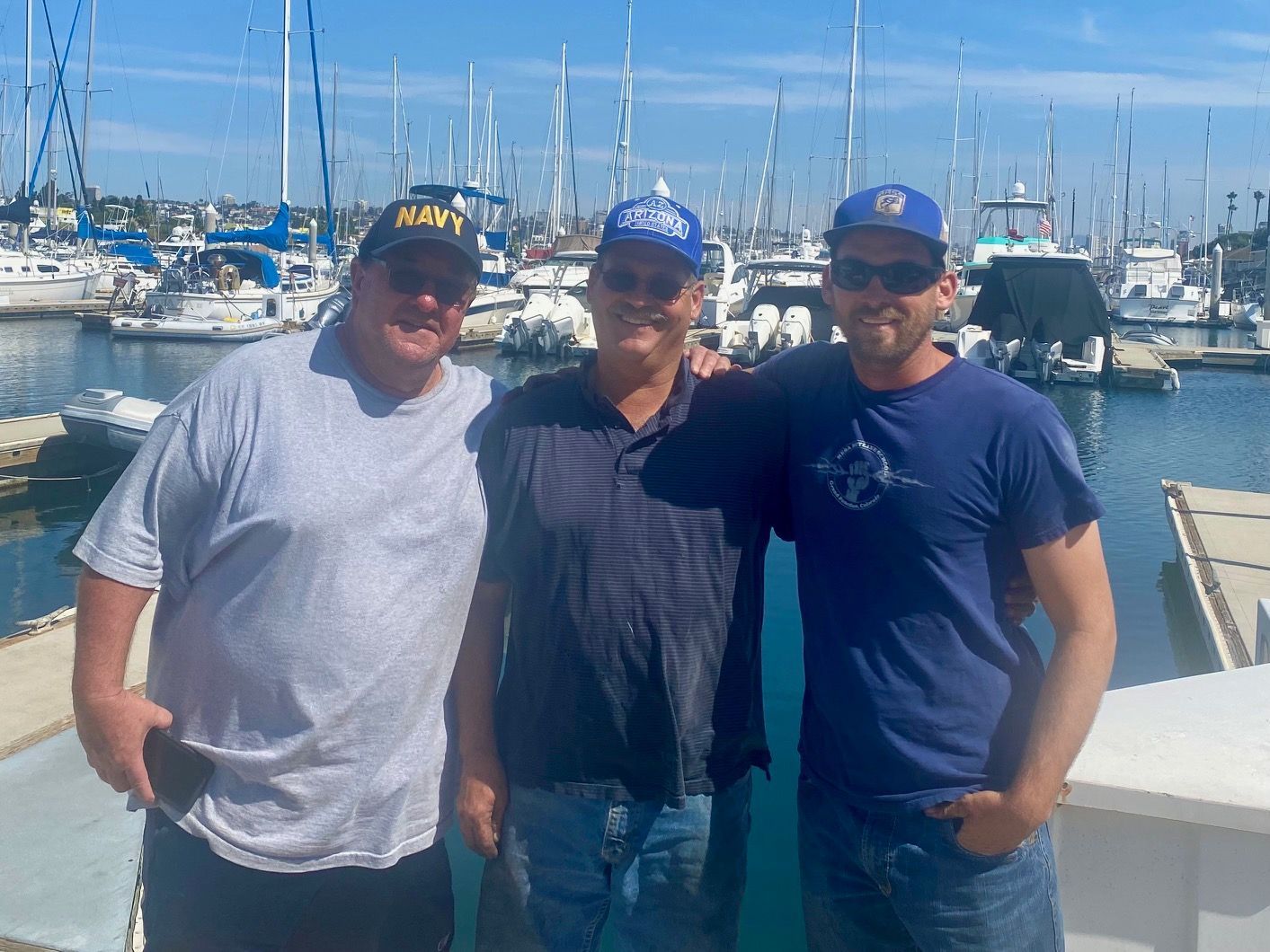 Three people wearing baseball caps stand side-by-side on a marina dock in front of a harbor full of moored sailboats.