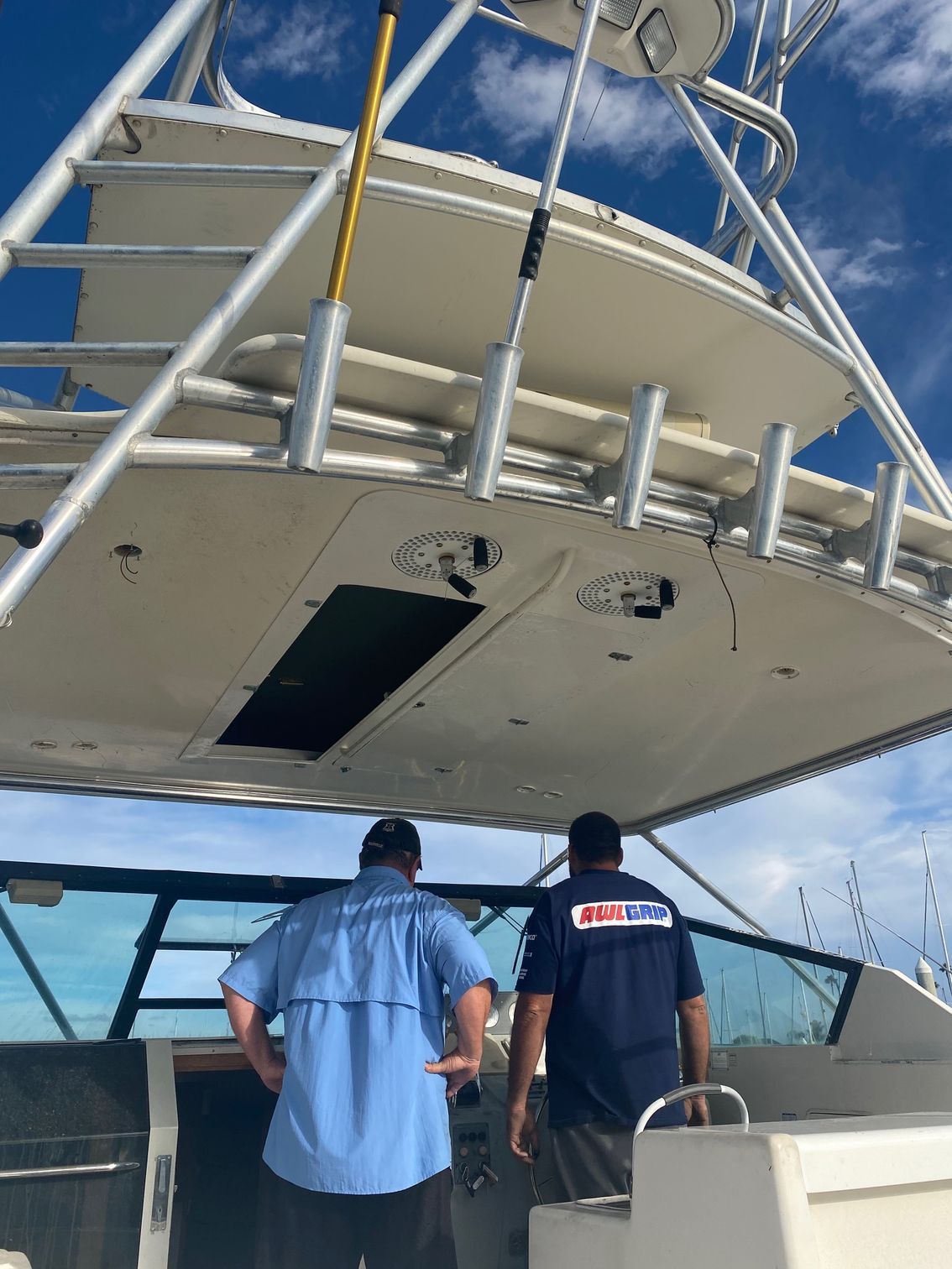 Two people stand at the helm of a boat under a metal hardtop structure with rod holders against a blue sky.