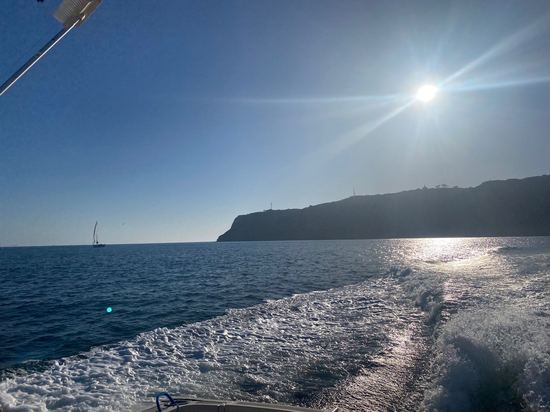 A view from a boat showing a white wake in the blue sea, leading toward a distant rocky coastline under a bright sun.