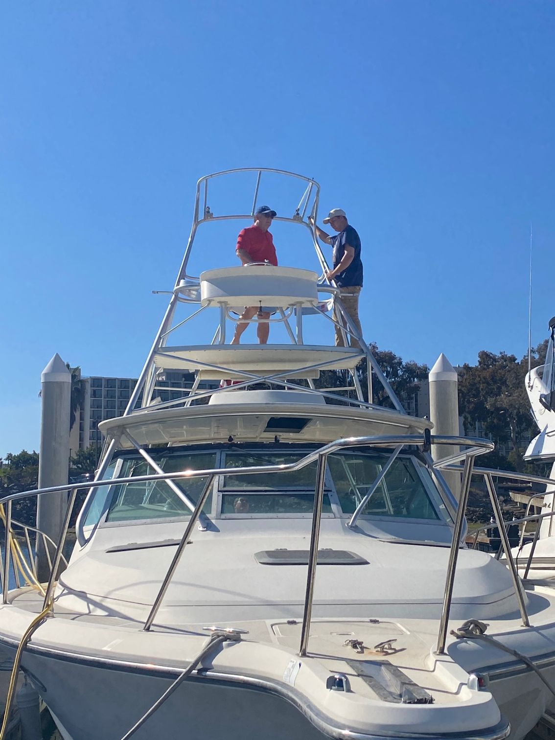 Two people stand on a metal boat tower frame at a marina on a sunny day.
