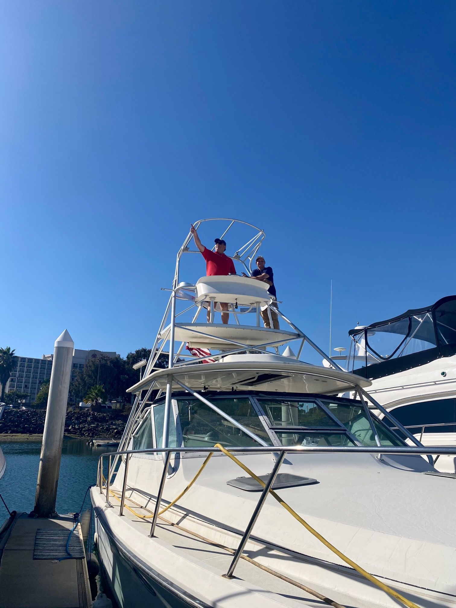 Two people stand on the upper deck of a white boat docked in a sunny harbor.