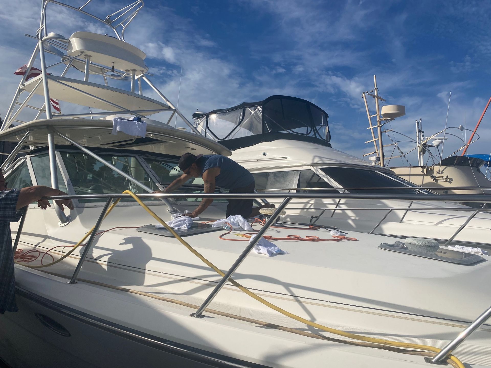 A person cleans the deck of a boat docked in a marina under a sunny blue sky.