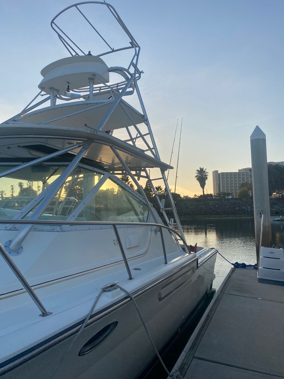 A white boat docked at a pier during sunset, with a tall metal tower and fishing poles visible against the sky.