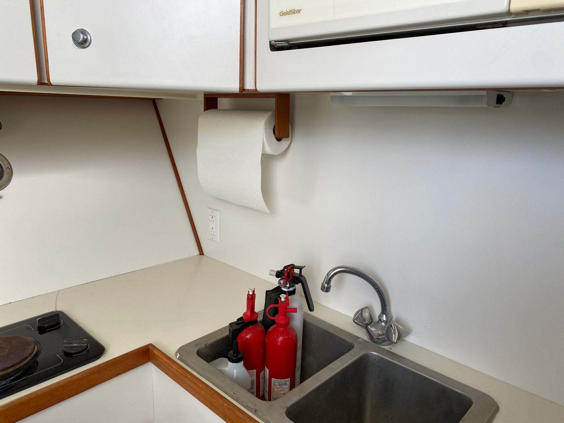 A kitchen sink on a boat with two red fire extinguishers sitting inside the basin, next to a stove and paper towels.