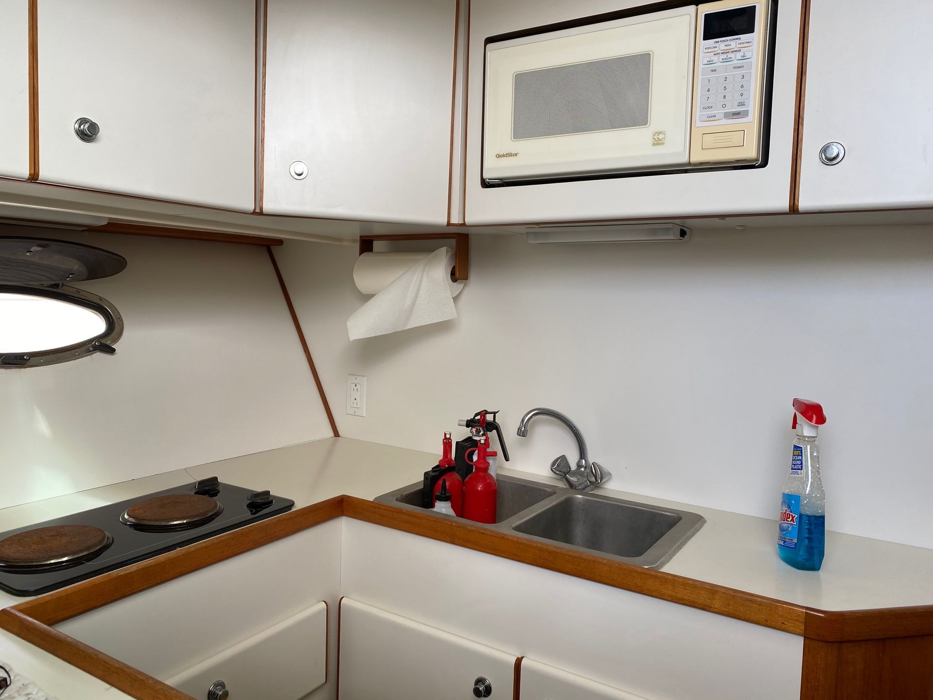 A compact galley kitchen on a boat, featuring a double sink, a two-burner stove, a wall-mounted microwave, and cabinetry.