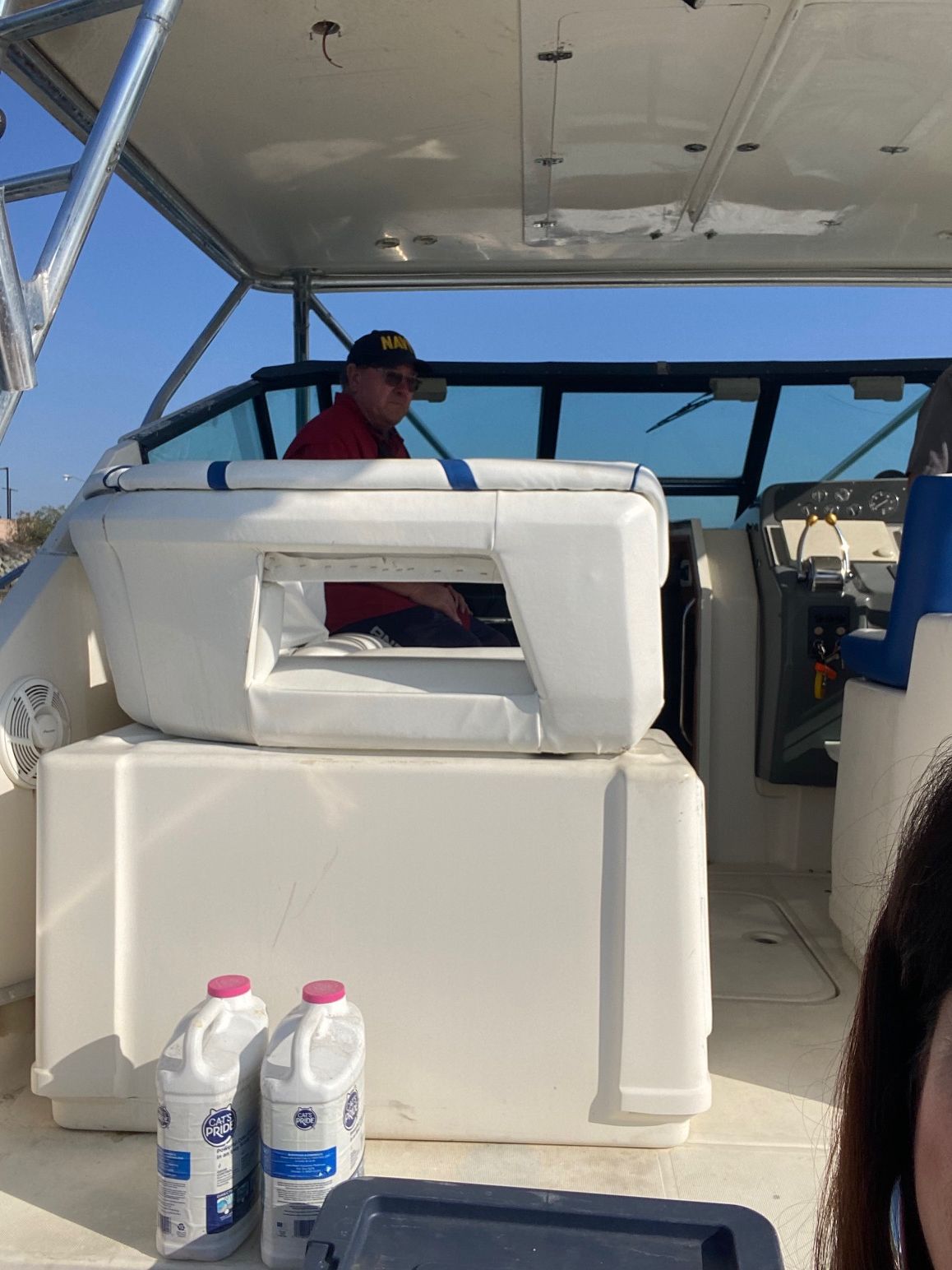 A person in a red shirt and cap sits at the helm of a boat, with two white plastic jugs in the foreground.