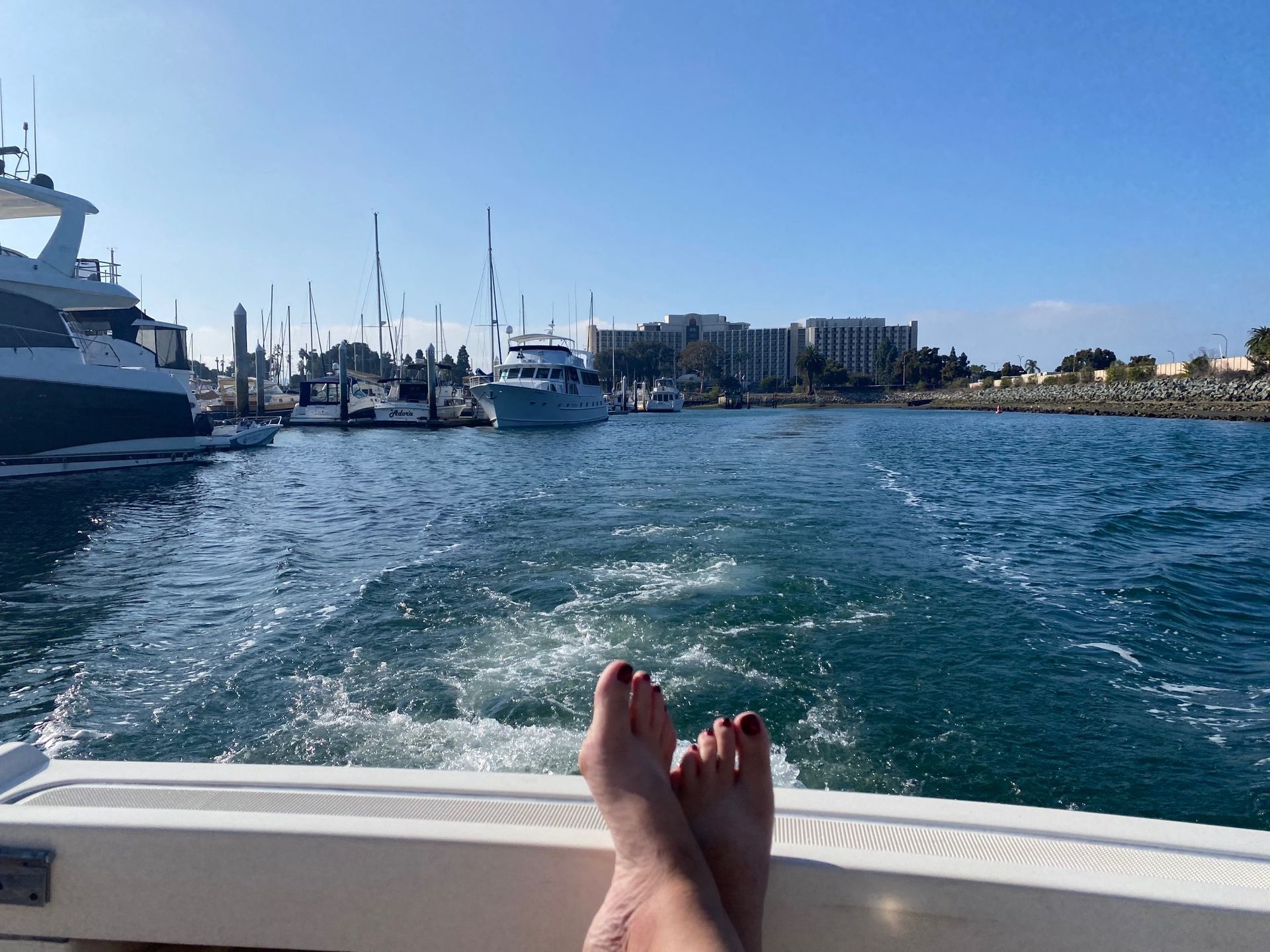 A person's feet resting on the deck of a boat moving through a marina toward a large building under a clear blue sky.