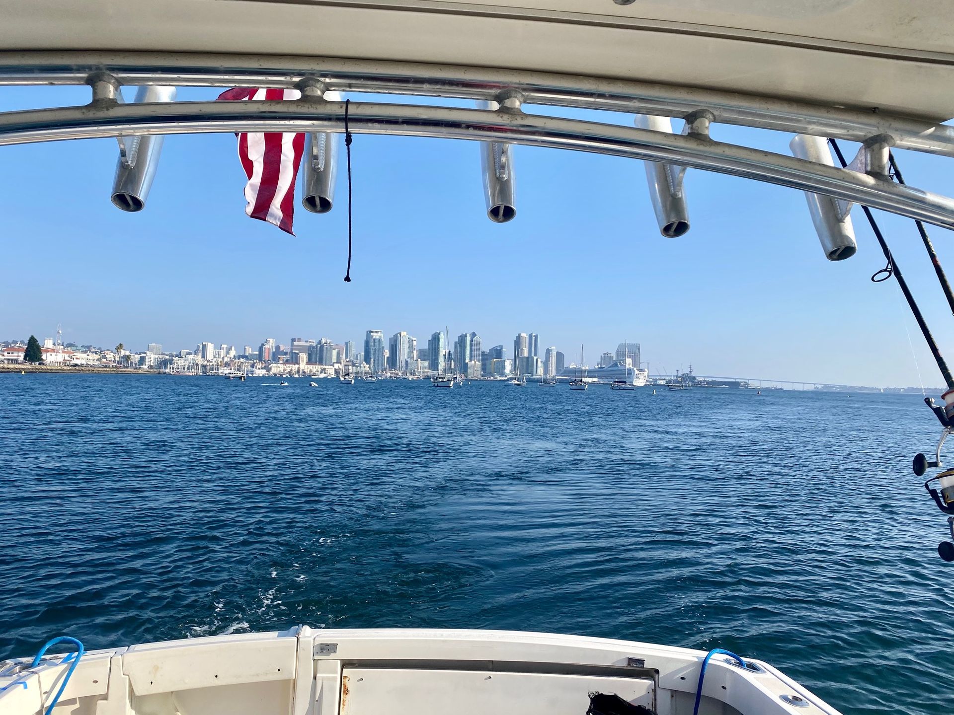 View of a city skyline across the water from the deck of a boat, framed by a metal rod rack holding fishing poles.