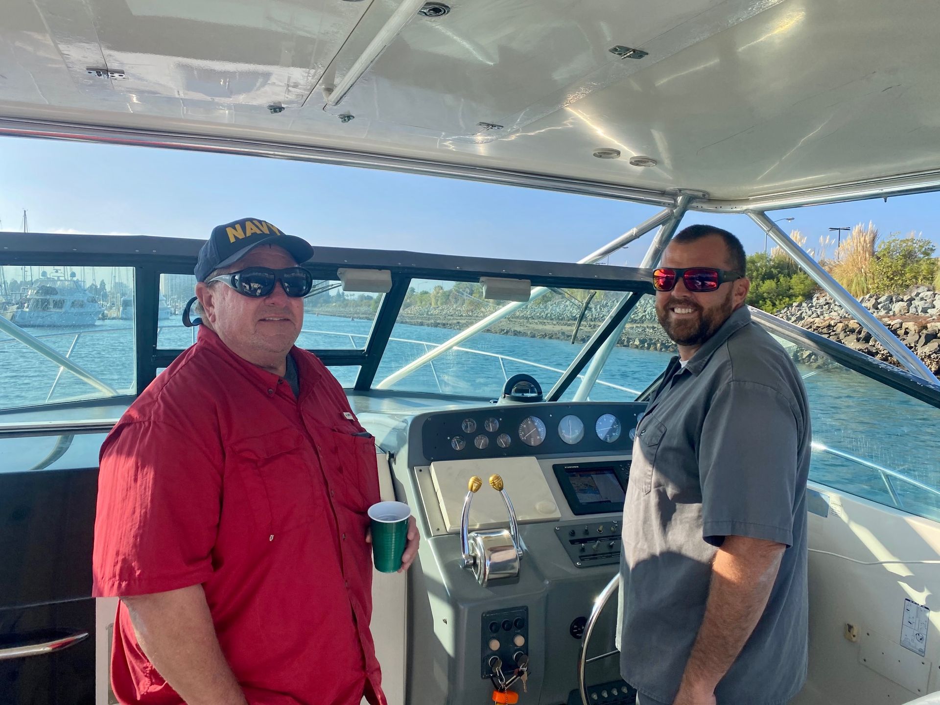 Two men stand on the bridge of a boat on a sunny day, smiling and wearing sunglasses.