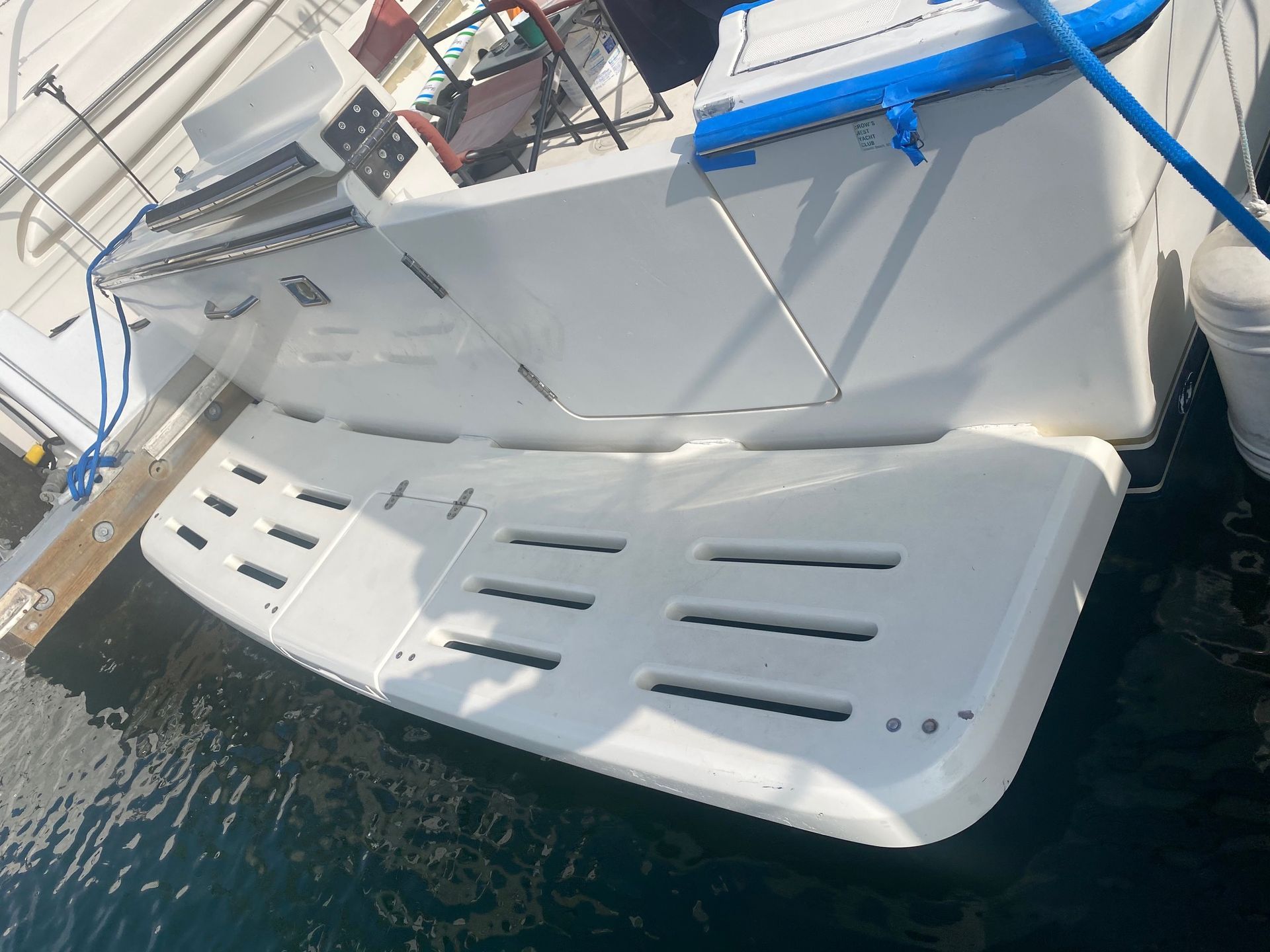 A white boat swim platform with slatted drainage, attached to the stern of a boat docked at a marina over dark water.
