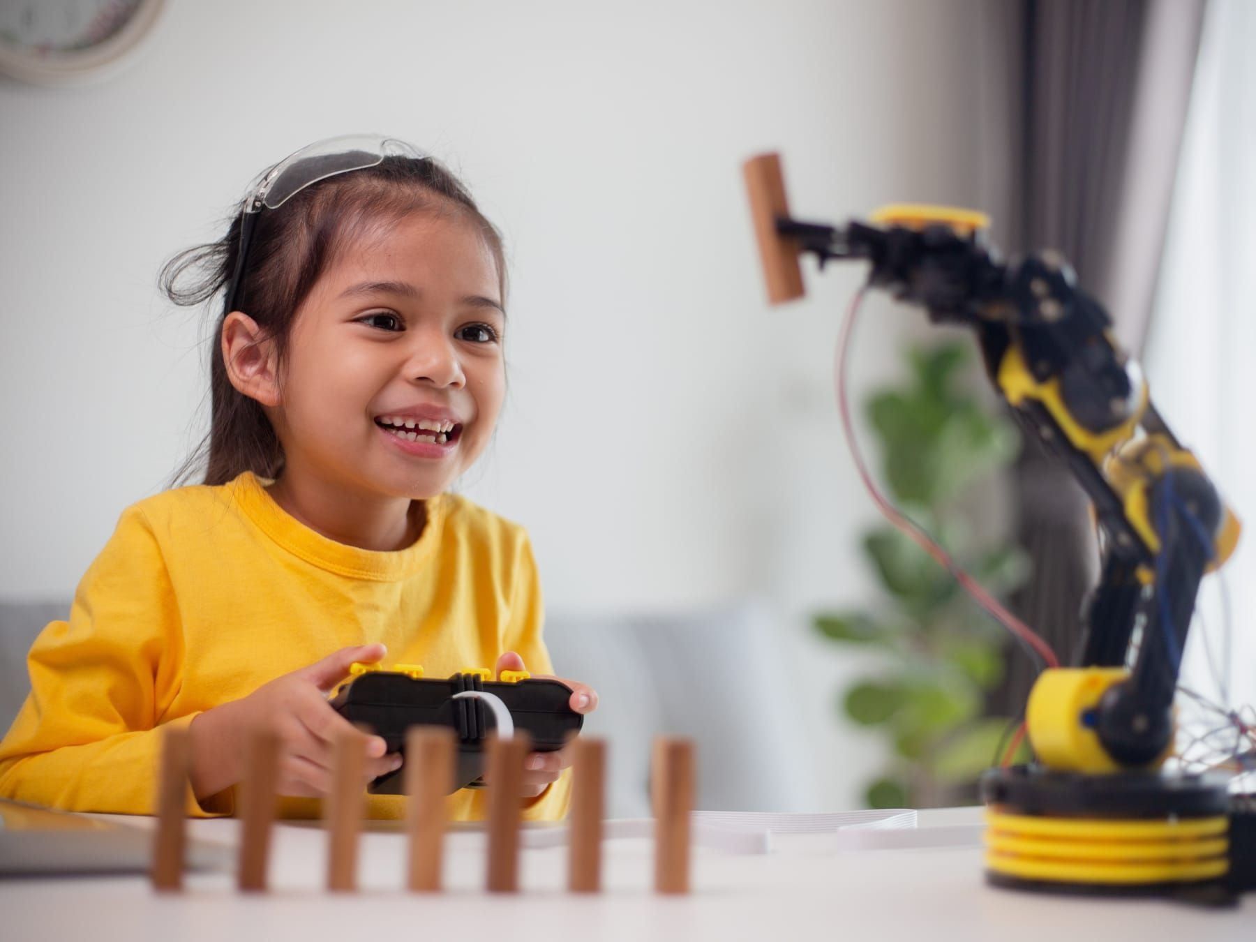 Girl smiling, controlling a robotic arm with a remote, arranging wooden blocks on a table.