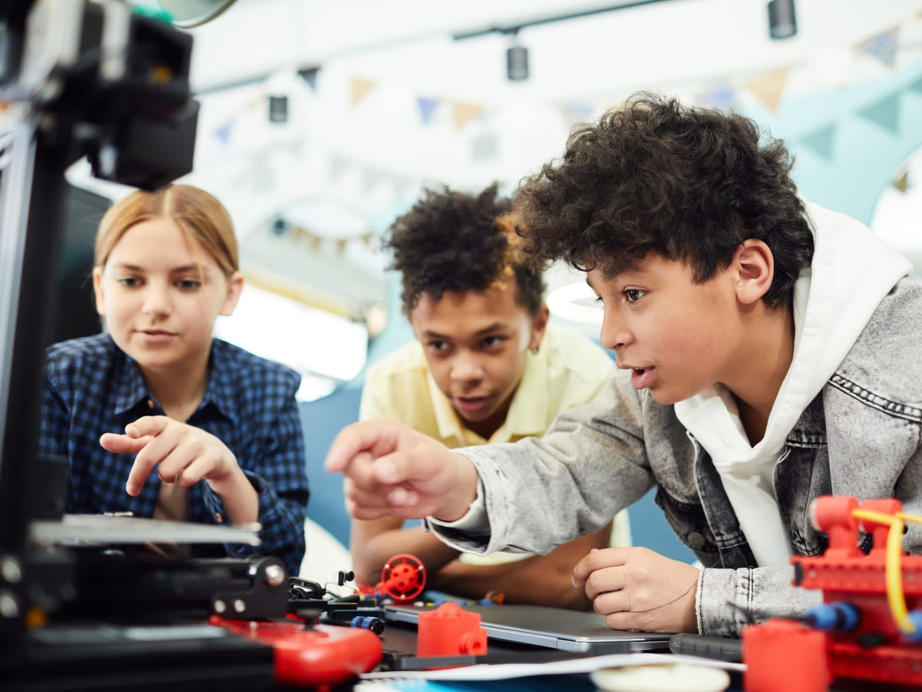 Three students examine a 3D printer. A boy points, while a girl and boy look on. Indoors with colorful accents.