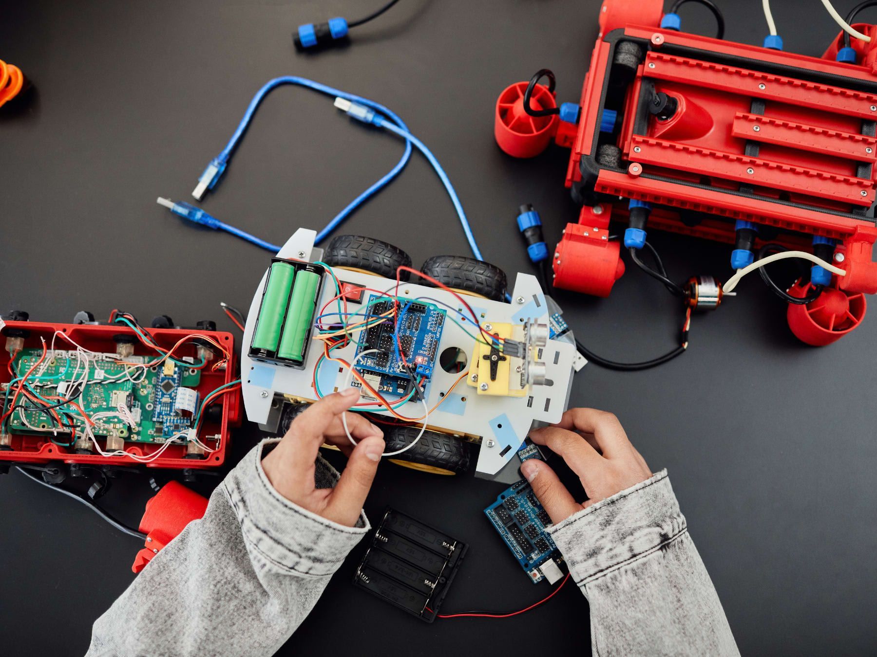 Hands assembling robots with wires, circuit boards, and batteries on a black surface.