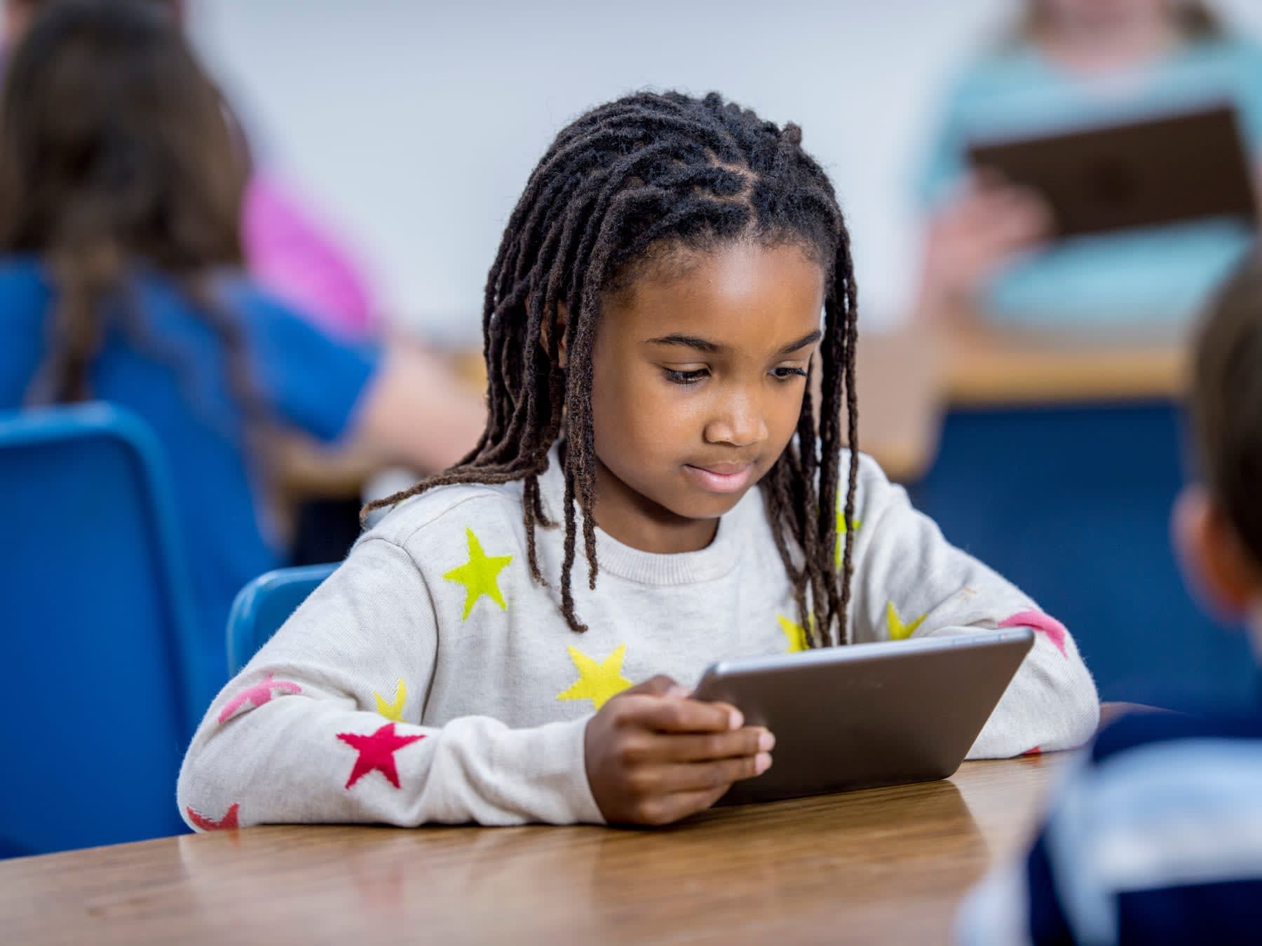 Young Black girl with dreadlocks using a tablet at a desk in a classroom.