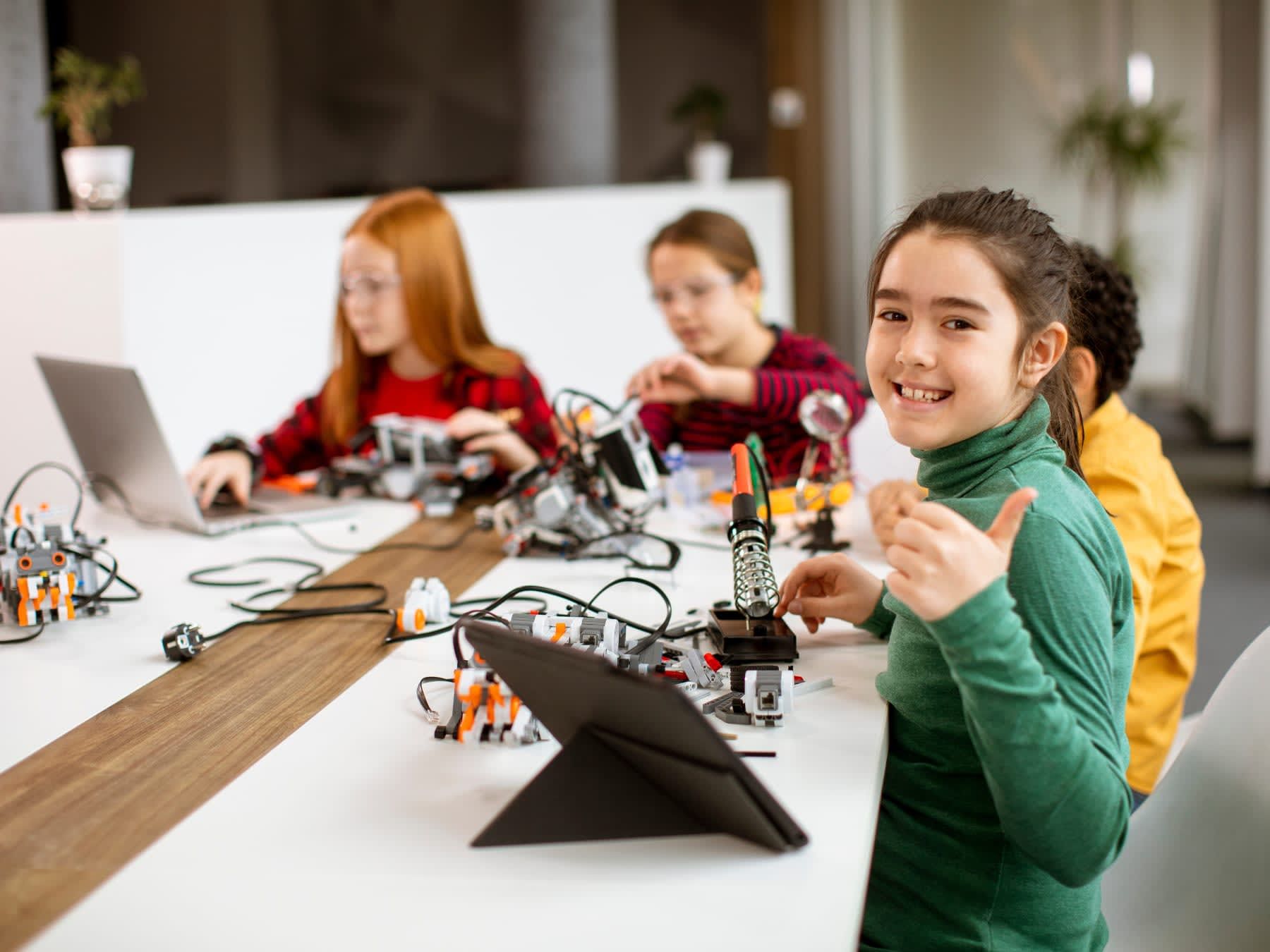 Group of children in robotics class, building robots at a table, one girl giving thumbs up.