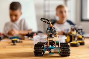 Boy holding a robot in a classroom; other children and adults in the background.