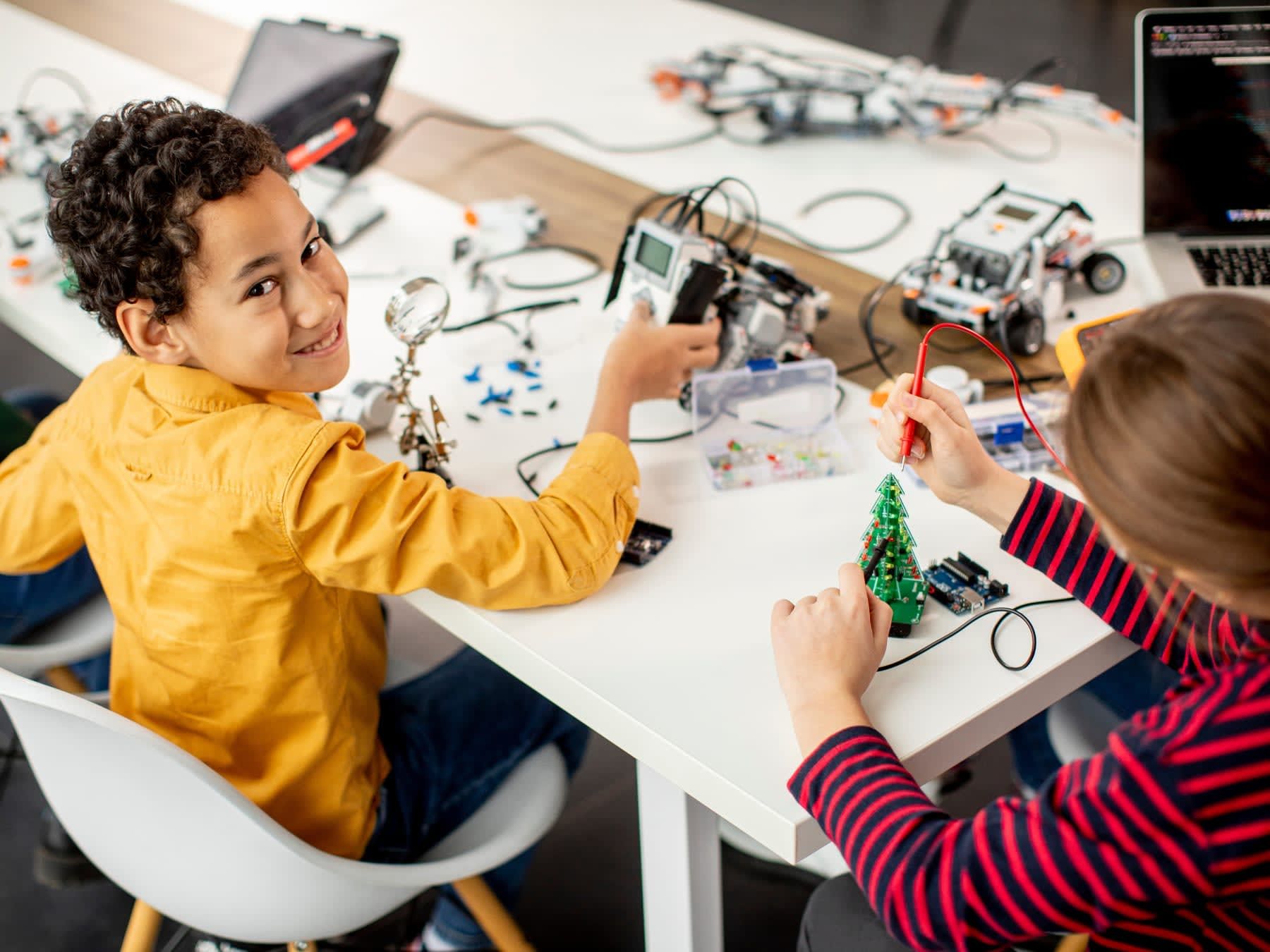 Two children building robots at a table, smiling. Bright room, robots, and tools.