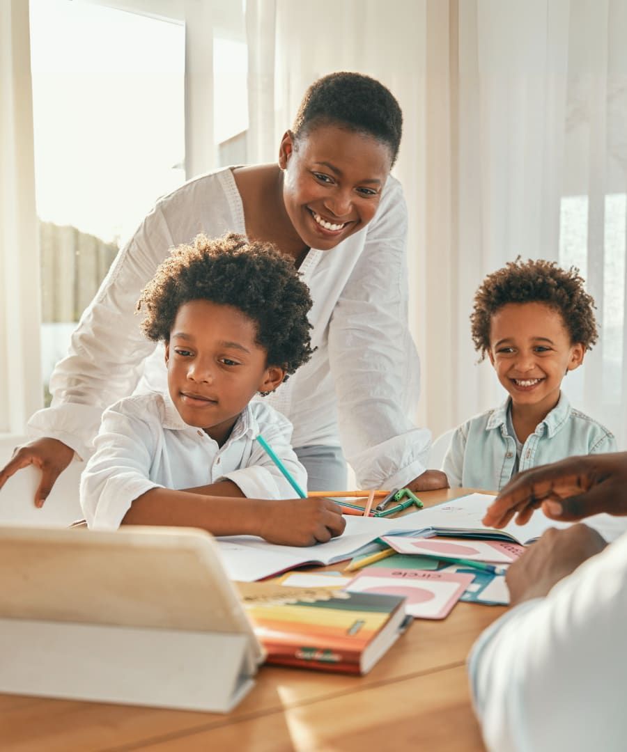 A smiling mother helps her two young sons with their schoolwork at a wooden table, bright setting.