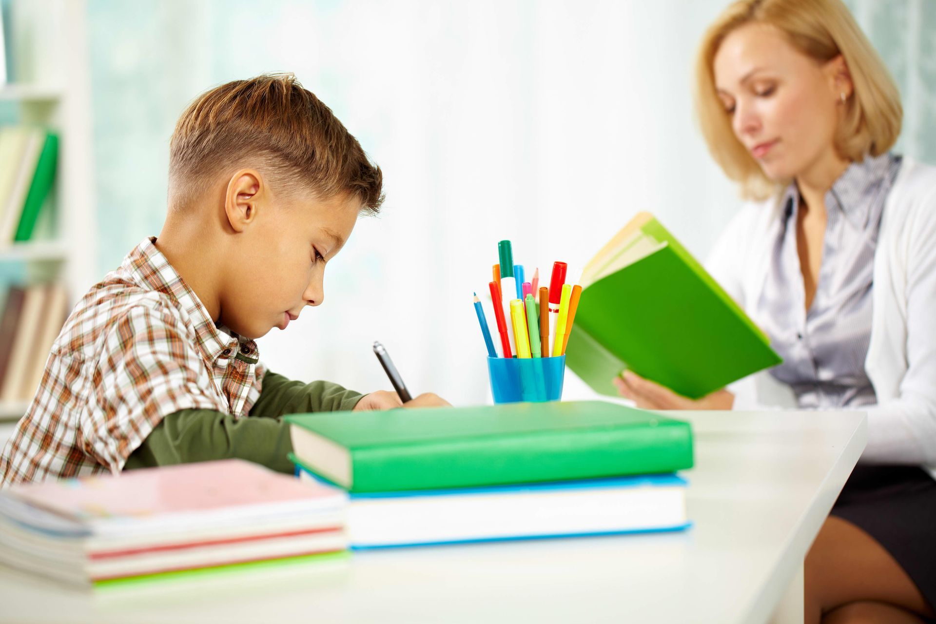 Boy writing at a desk, being tutored by a woman holding a book.