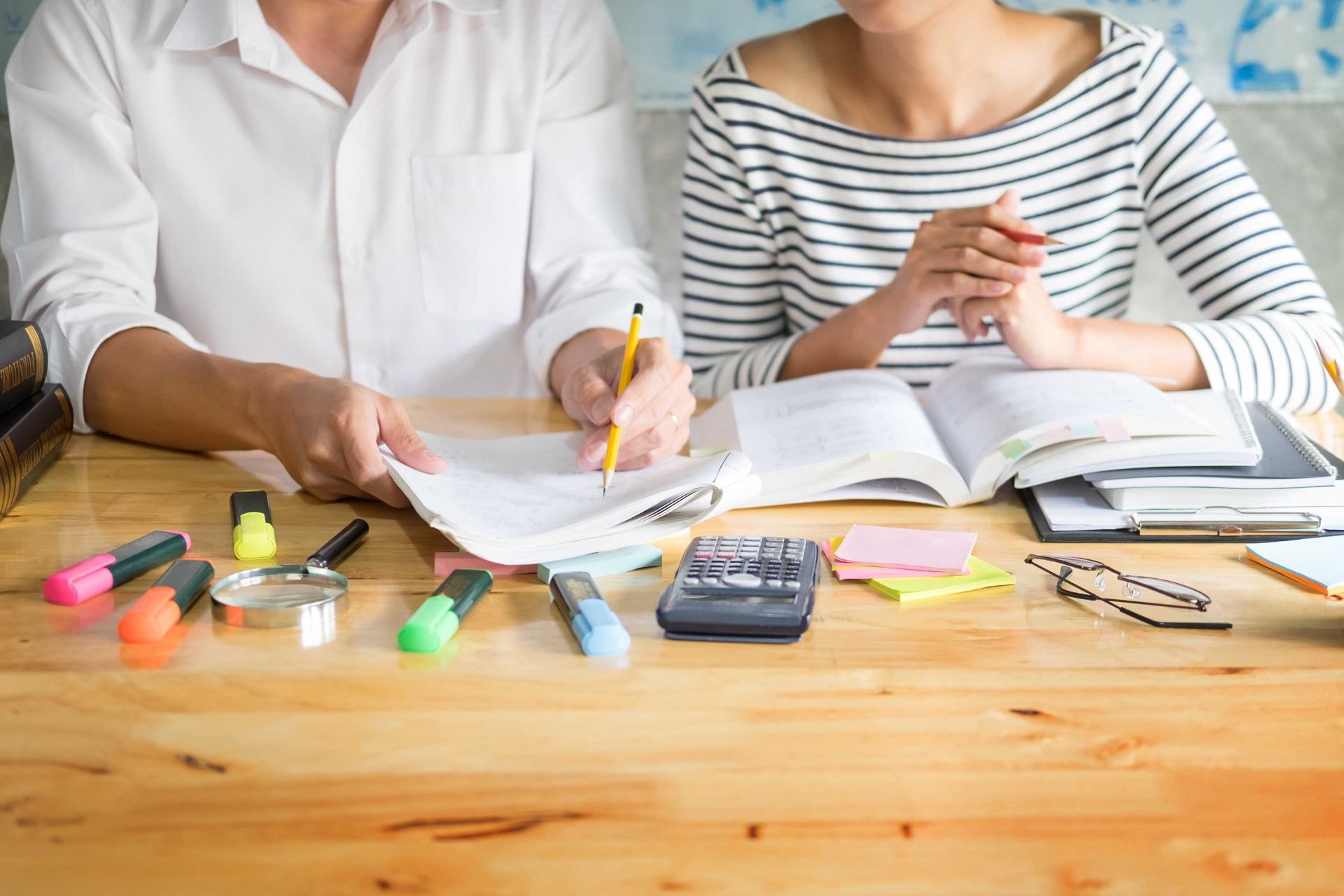 Two people studying together at a wooden table with books, calculator, and highlighters.