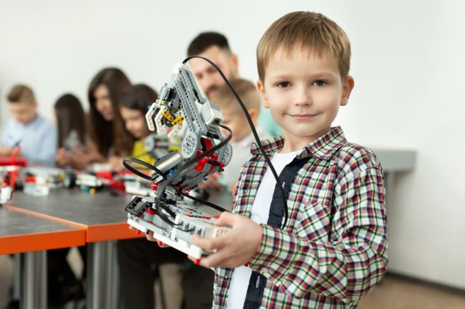 Boy holding a Lego robot, smiling at the camera in a classroom. Other kids and adults in background.