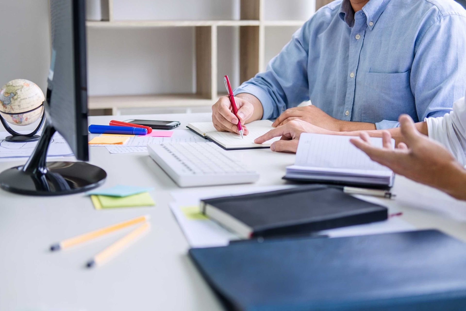 Two people collaborating at a desk, one writing in a notebook, while the other gestures and speaks.