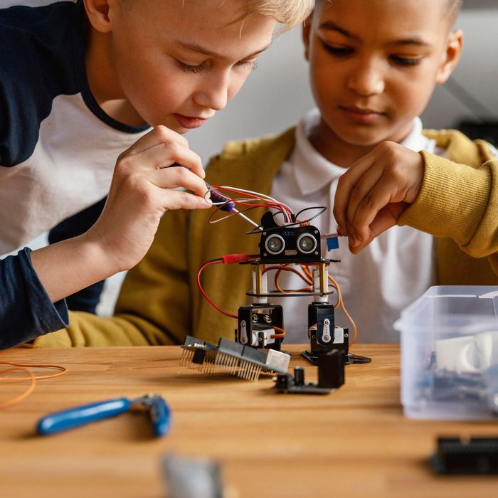 Robot arm made of LEGO pieces in classroom, reaching for a metal cylinder.