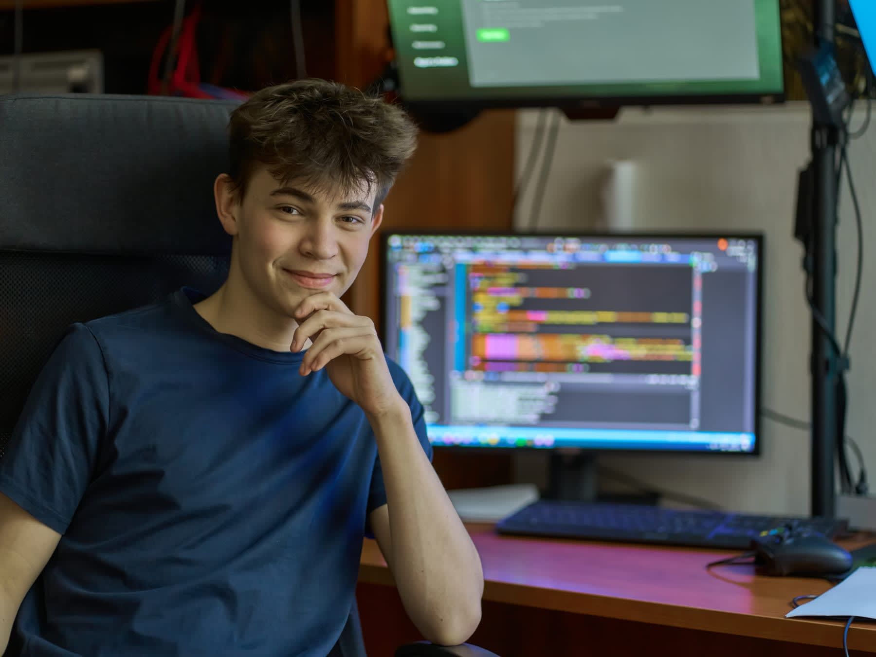 Smiling young man with brown hair, seated at a desk with computer monitors showing code.