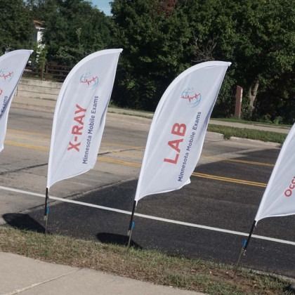 White teardrop flags on a road with "X-Ray" and "Lab" printed on them, promoting a Minnesota mobile event.