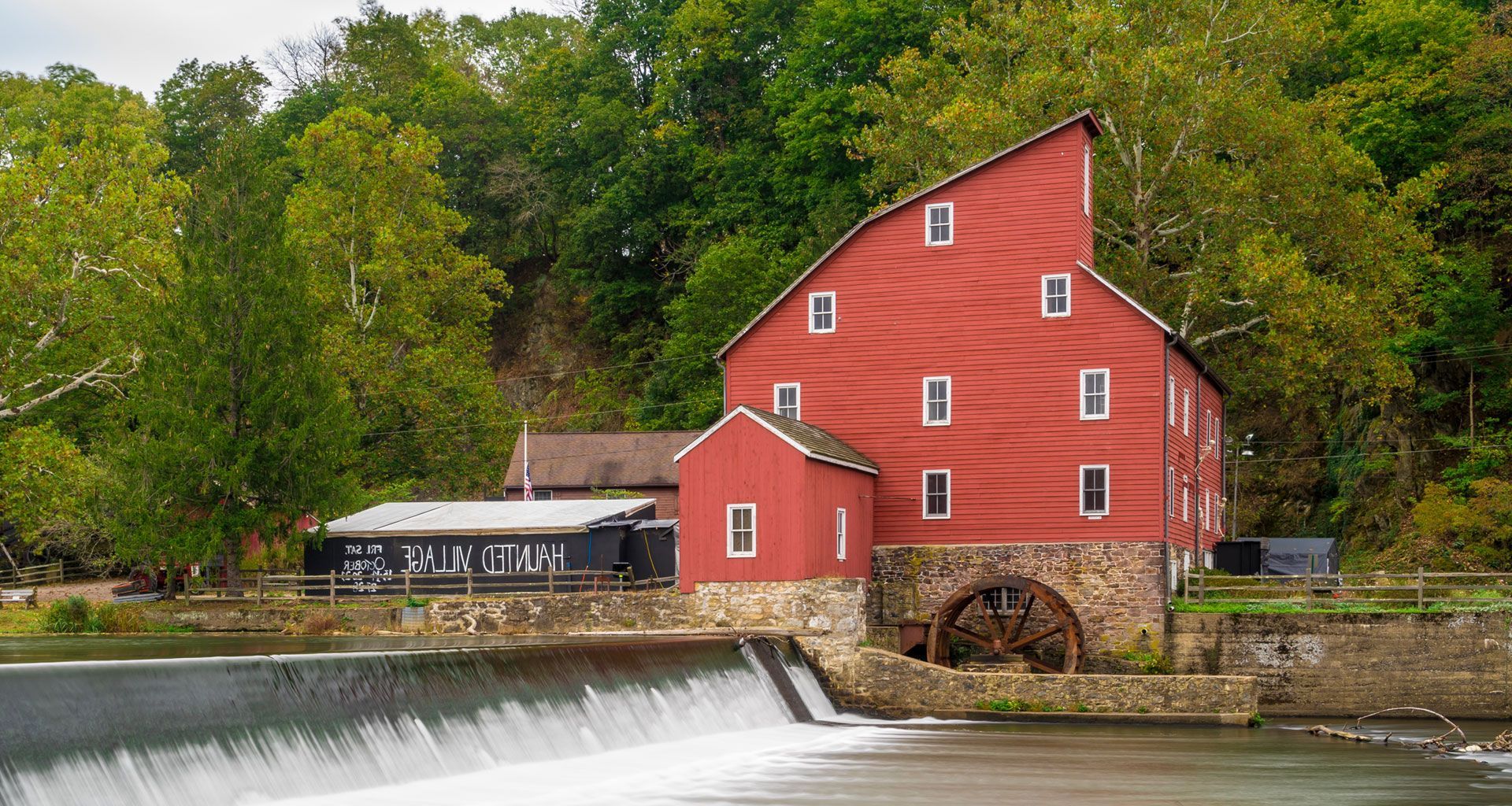 A red barn is sitting next to a river with a water wheel in front of it.