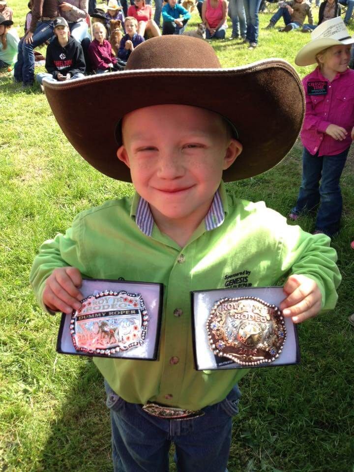 Smiling young boy in cowboy hat holds two belt buckles outdoors. He wears a green shirt and jeans | Genesis Automotive and RV Repair