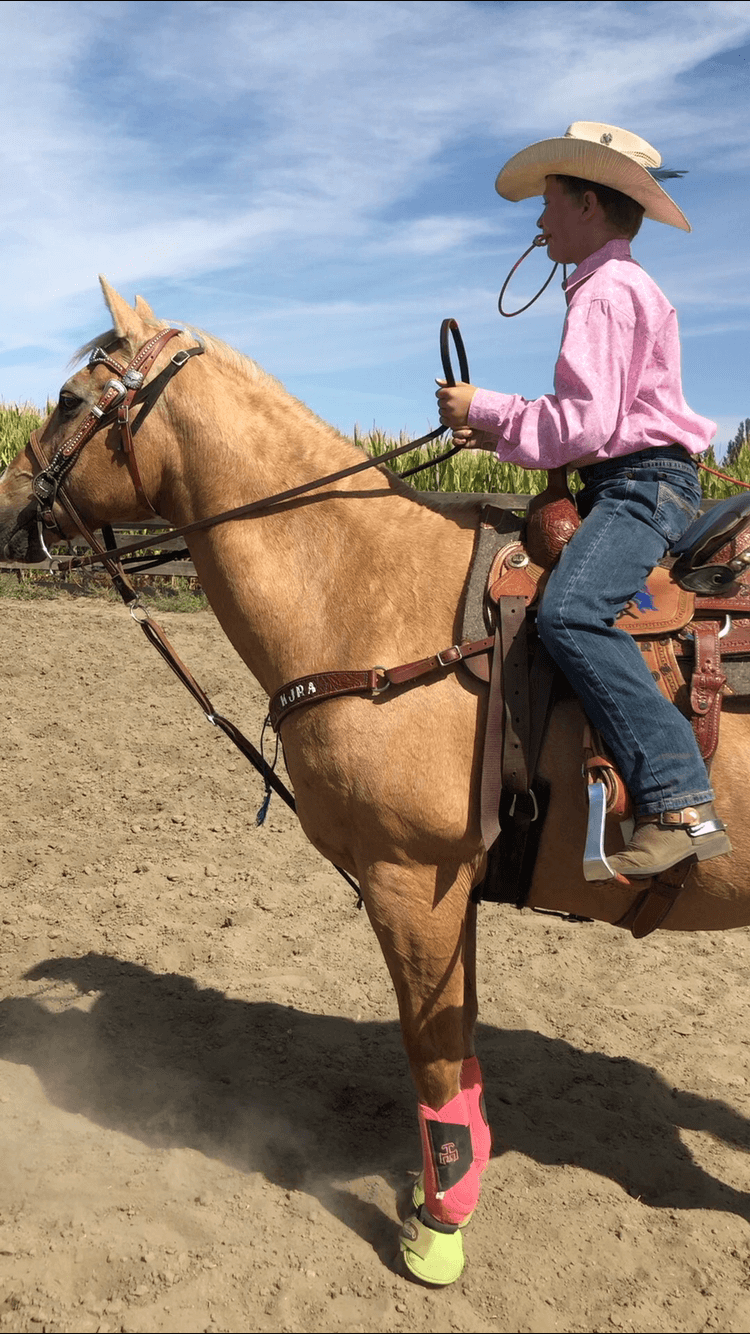 Boy in cowboy hat on a tan horse in an outdoor arena. Pink shirt, blue jeans, and pink leg wraps | Genesis Automotive and RV Repair