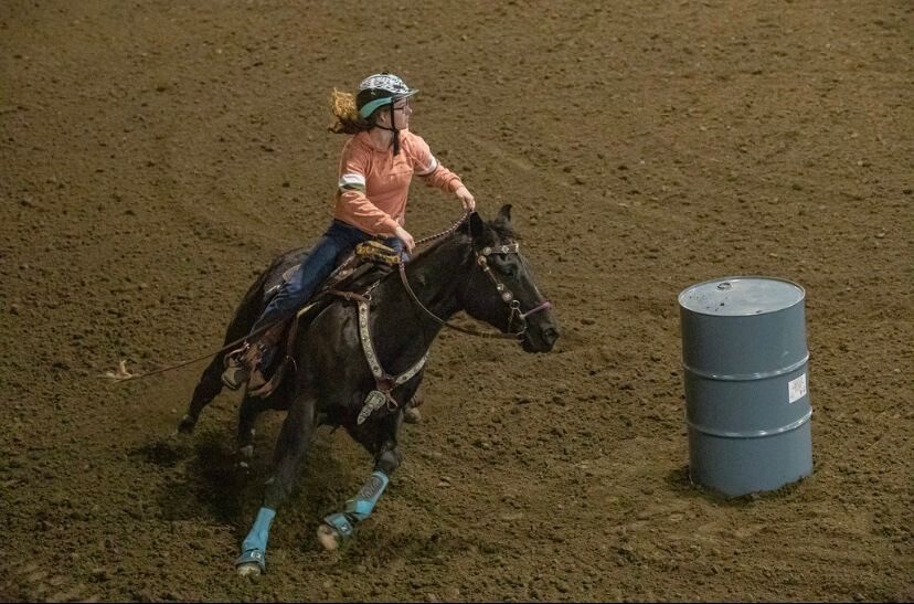 A cowgirl on a dark horse rounds a barrel in a dirt arena. The horse has blue leg wraps | Genesis Automotive and RV Repair