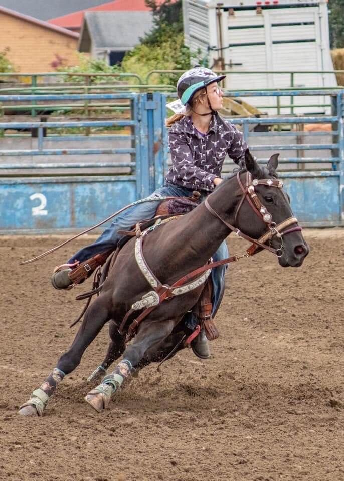A young woman on a dark horse, barrel racing in a rodeo, wearing a helmet and purple shirt | Genesis Automotive and RV Repair