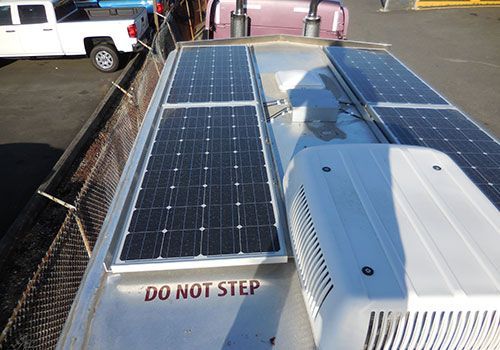 Solar panels mounted on the roof of a vehicle, near an air conditioning unit. Warning sign says,