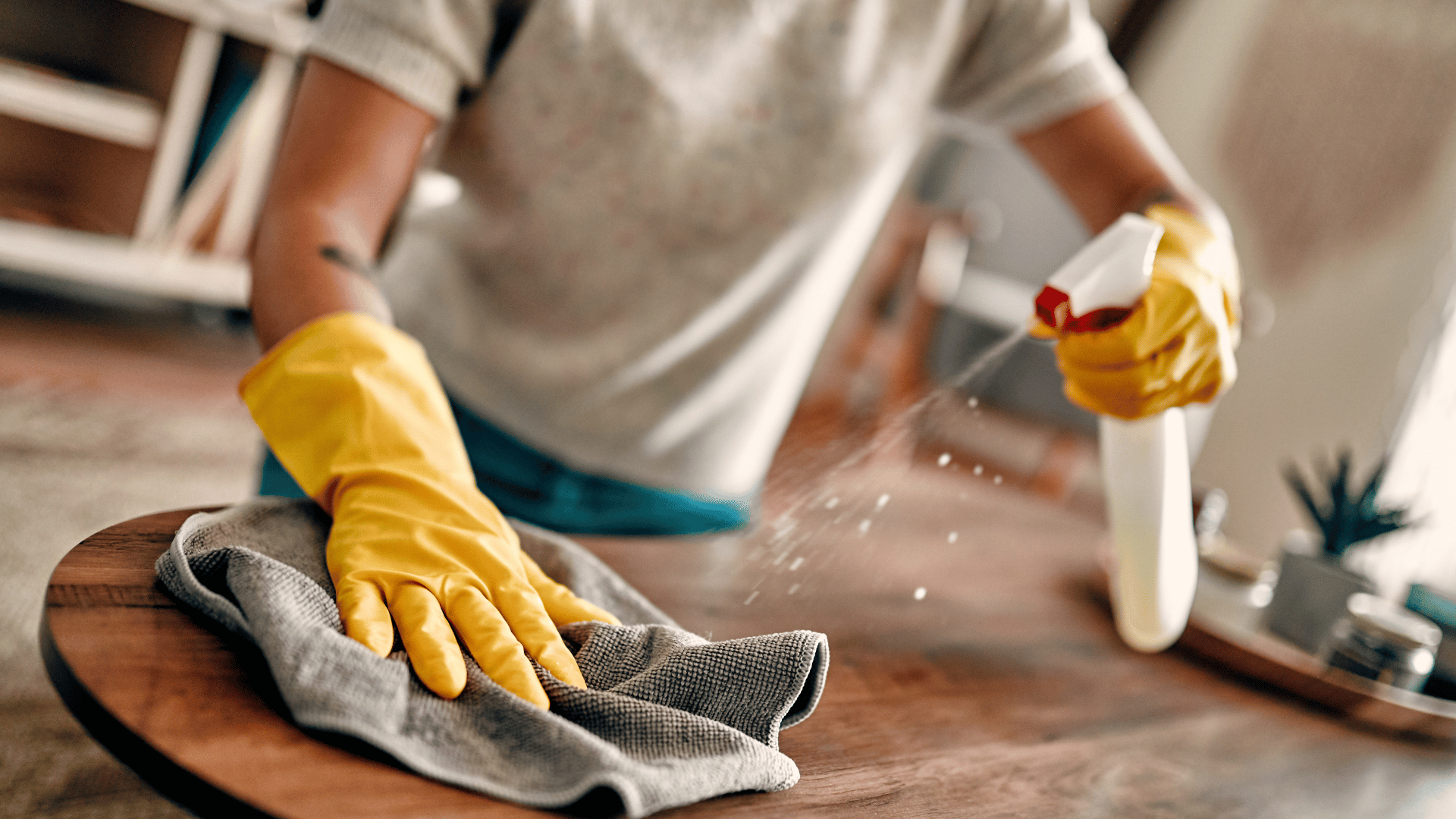 A person wearing yellow gloves is cleaning a wooden table with a cloth and spray bottle.