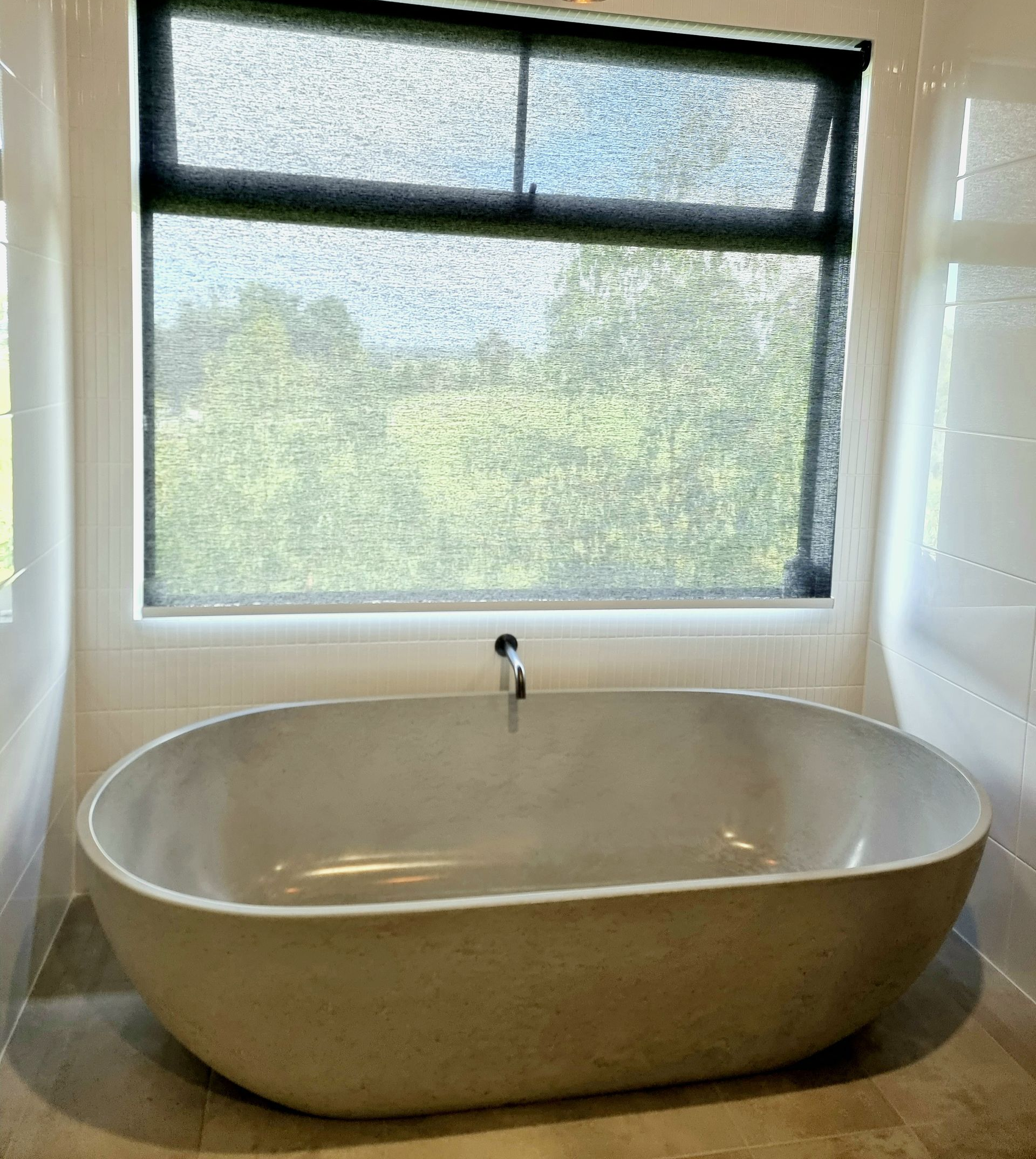 Oval bathtub under a window with a partially drawn black roller shade. Green scenery visible. — Glenn Koek - Shutters - Curtains - Blinds in West Wollongong, NSW