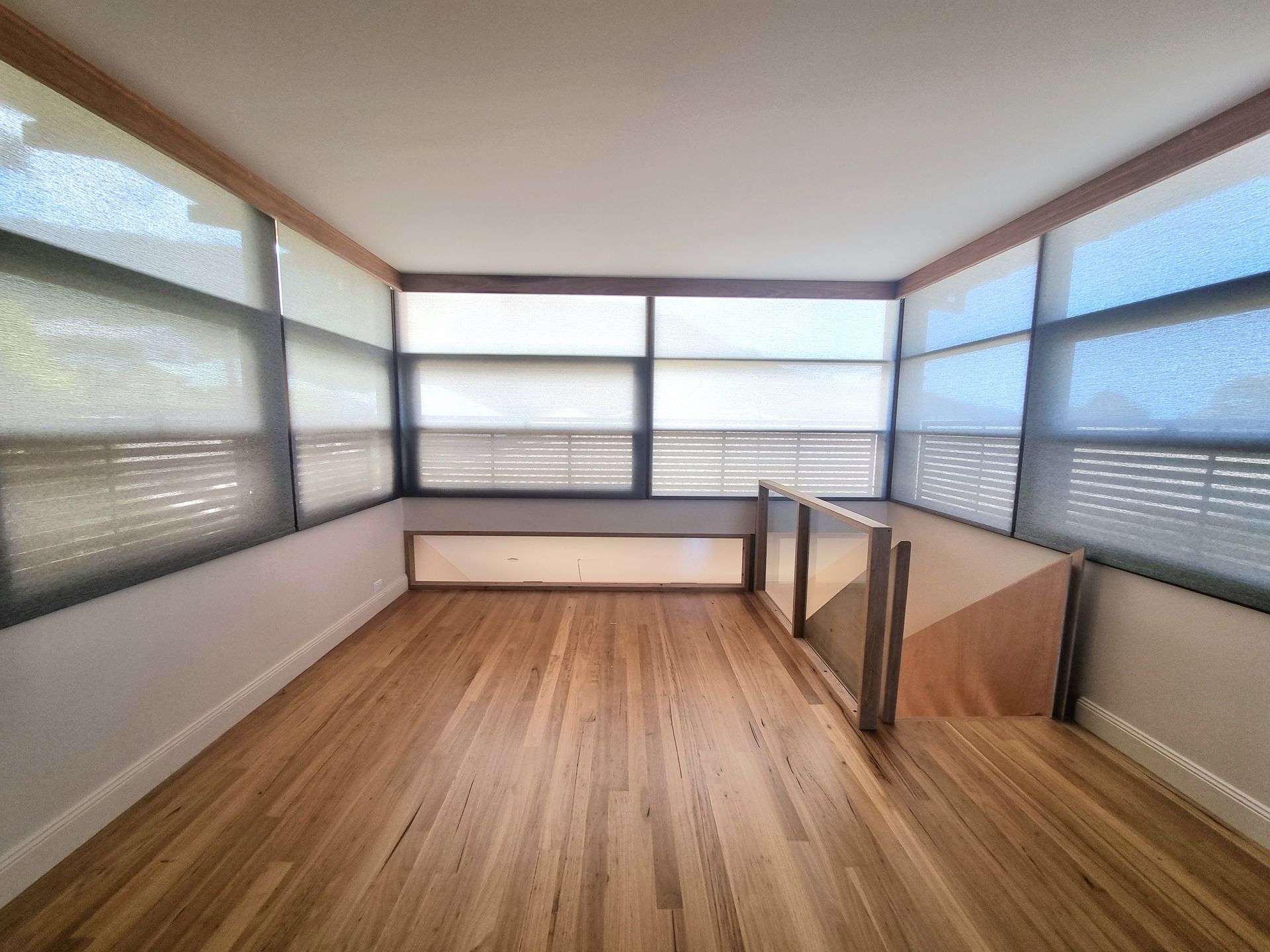 Wooden-floored interior with multiple windows covered by blinds; a short, wooden railing. — Glenn Koek - Shutters - Curtains - Blinds in West Wollongong, NSW