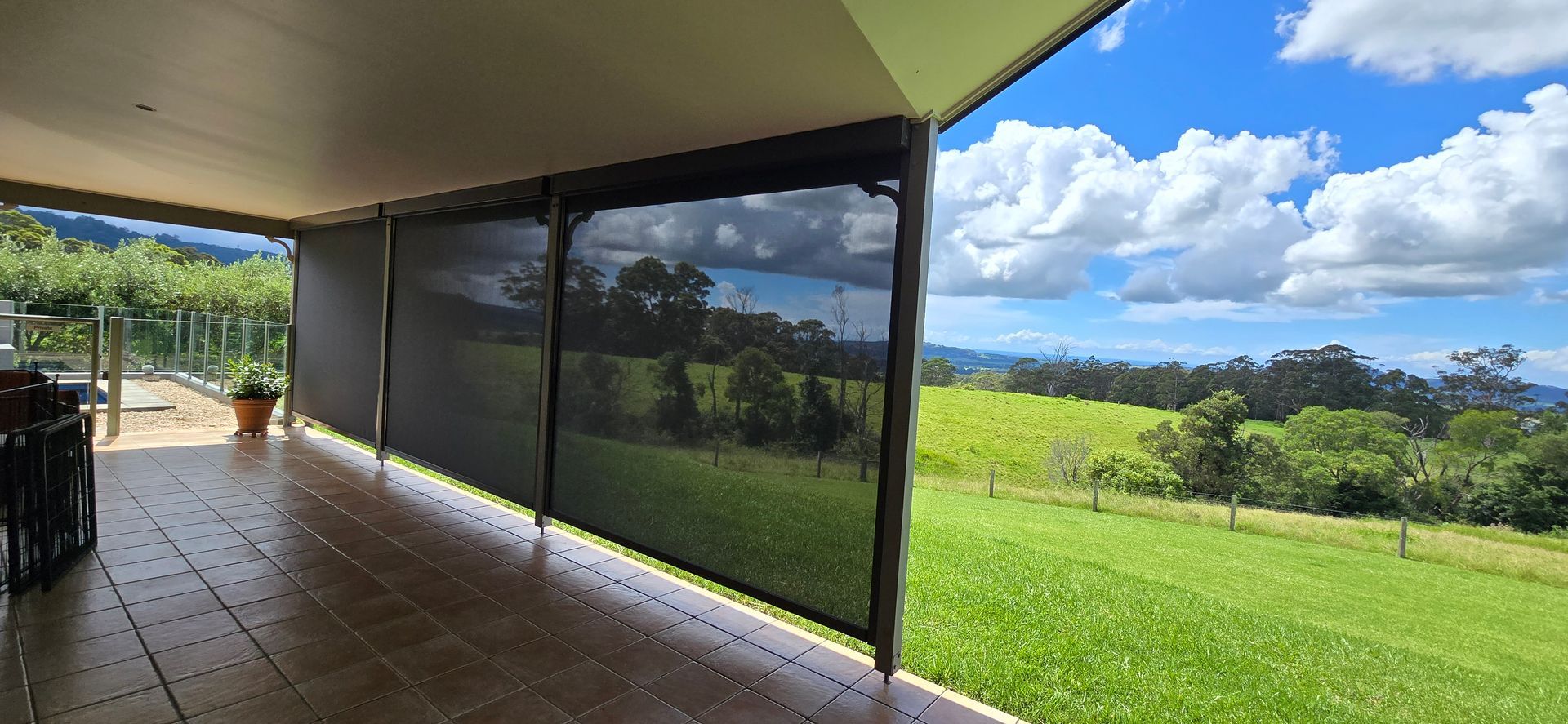 A covered patio with lowered black shades overlooking a green field under a blue sky with clouds. — Glenn Koek - Shutters - Curtains - Blinds in West Wollongong, NSW