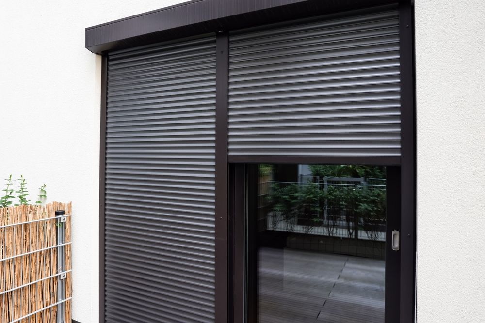 Exterior view of a dark-coloured roller shutter covering a door, partially open to reveal a balcony. — Glenn Koek - Shutters - Curtains - Blinds in West Wollongong, NSW