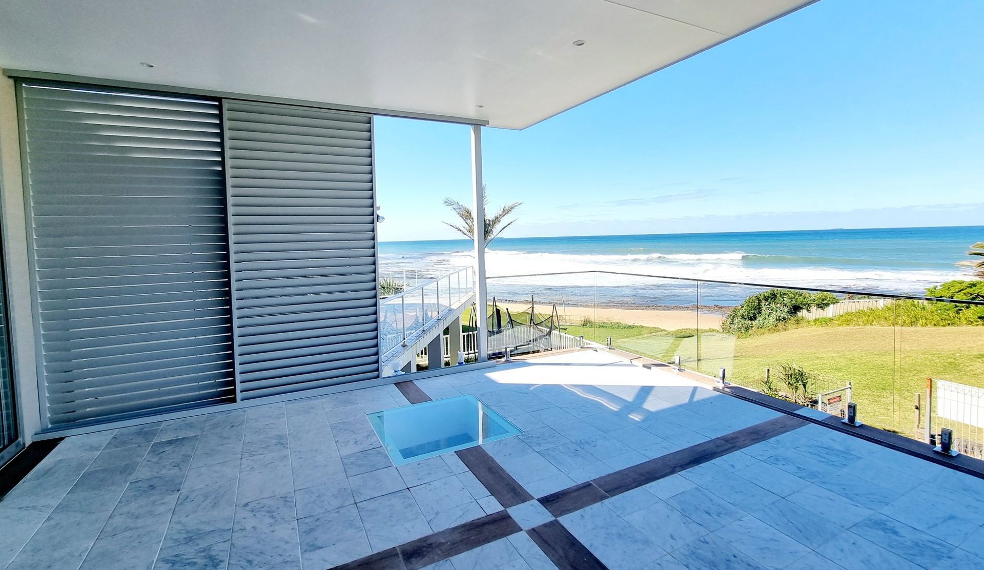 Oceanfront balcony with a view of the beach and ocean.  Sliding door, tiled floor, and blue sky. — Glenn Koek - Shutters - Curtains - Blinds in West Wollongong, NSW