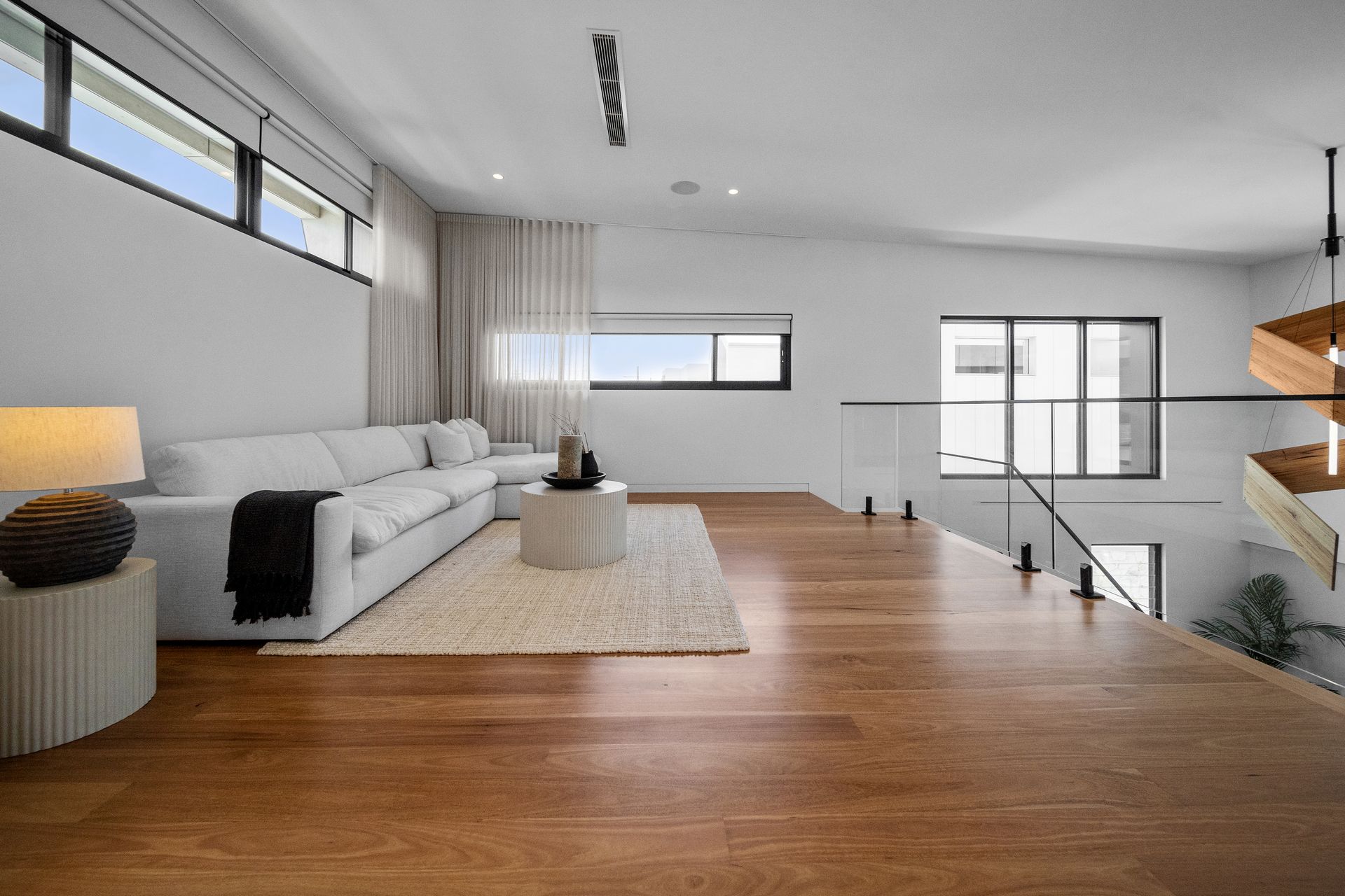 Modern living room with white sofa, rug, and wooden floors. Glass railing overlooks a lower level. — Glenn Koek - Shutters - Curtains - Blinds in West Wollongong, NSW