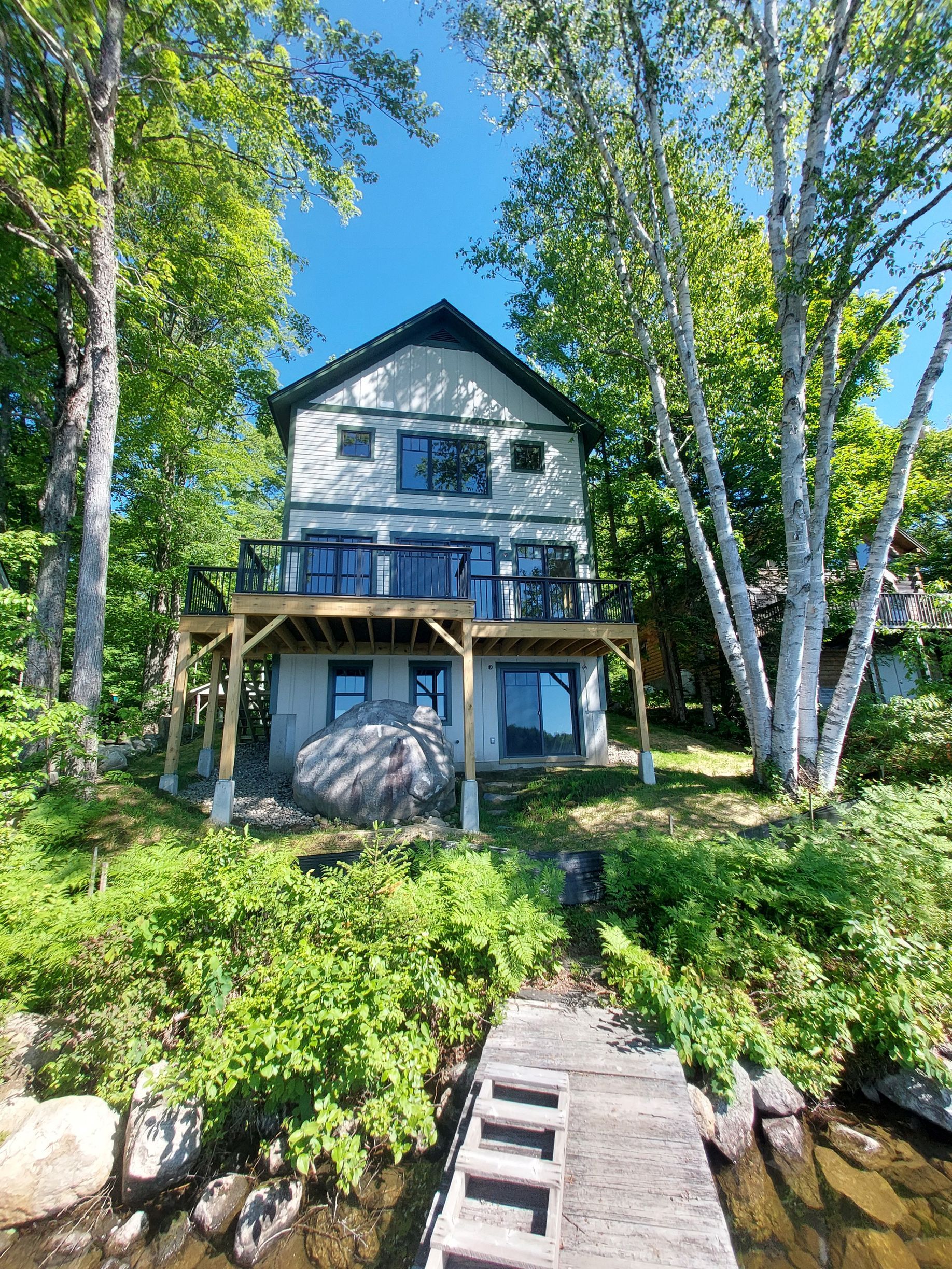 Blue lakeside house with porch, framed by tall trees and greenery, beside a small dock and rocky shoreline