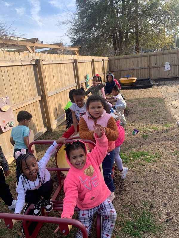 A group of children plays together on a red seesaw in a fenced outdoor playground.