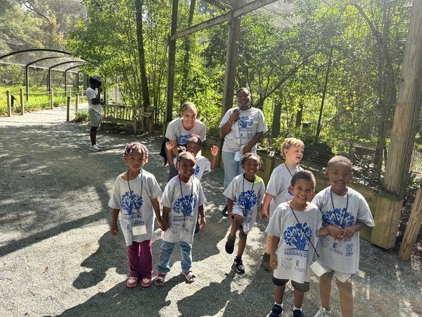 A group of children and adults wearing matching event T-shirts pose together on a gravel path at an outdoor park.