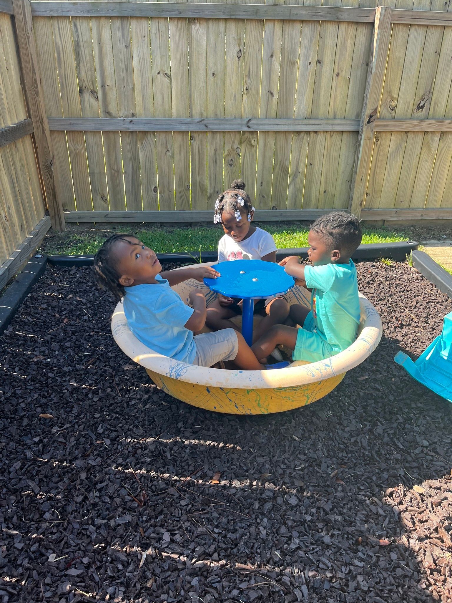 Three children sitting inside a yellow spinning playground toy on a wood chip surface near a wooden fence.
