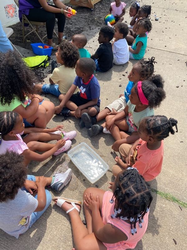 A group of children sits on pavement in a circle around a container of water, participating in an outdoor activity.