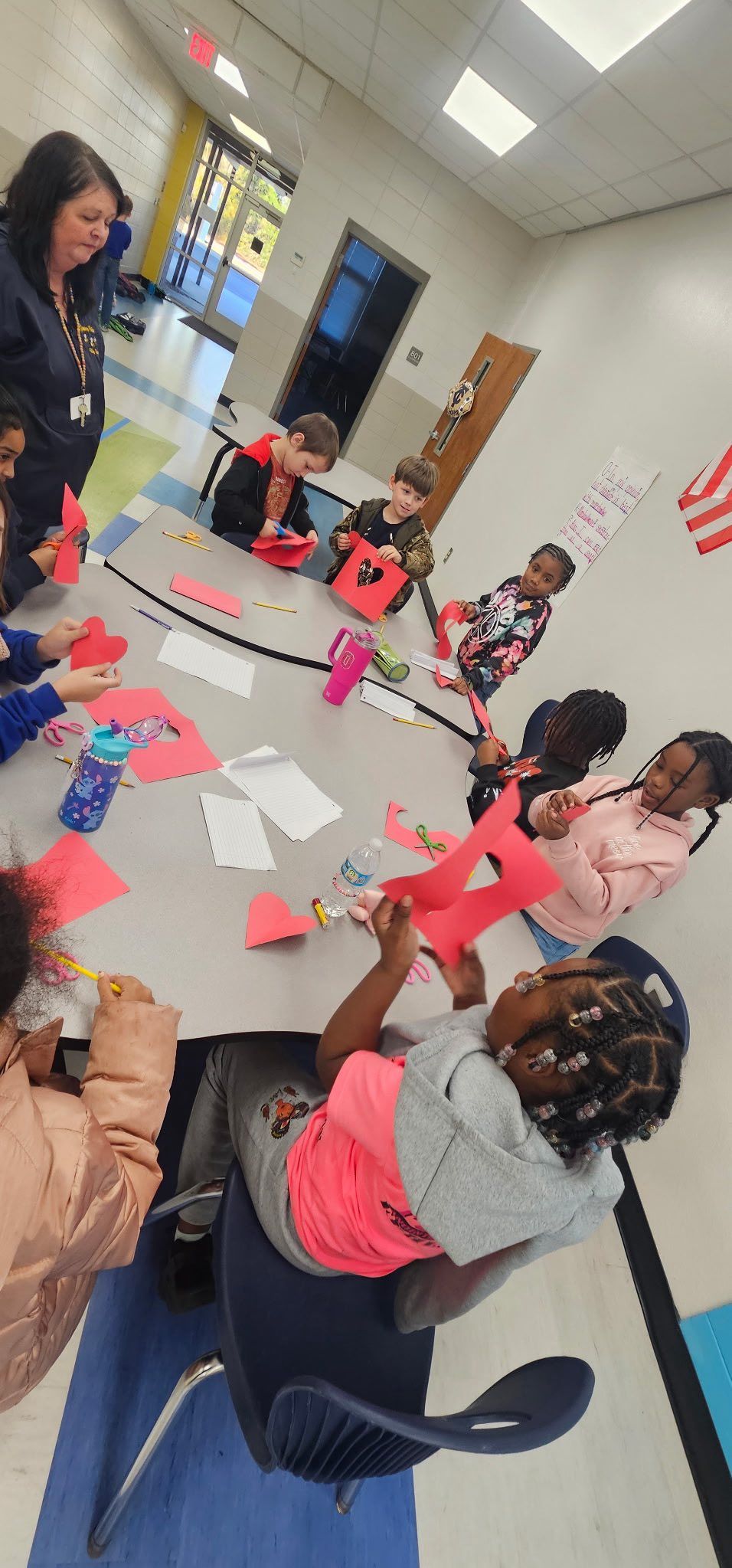 A teacher and students work on paper crafts at a large table in a bright classroom.