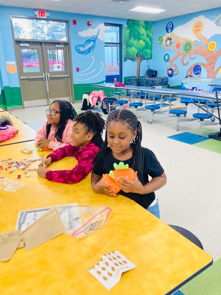 Three children sit at a bright yellow table in a classroom with colorful wall murals, working on a craft activity.