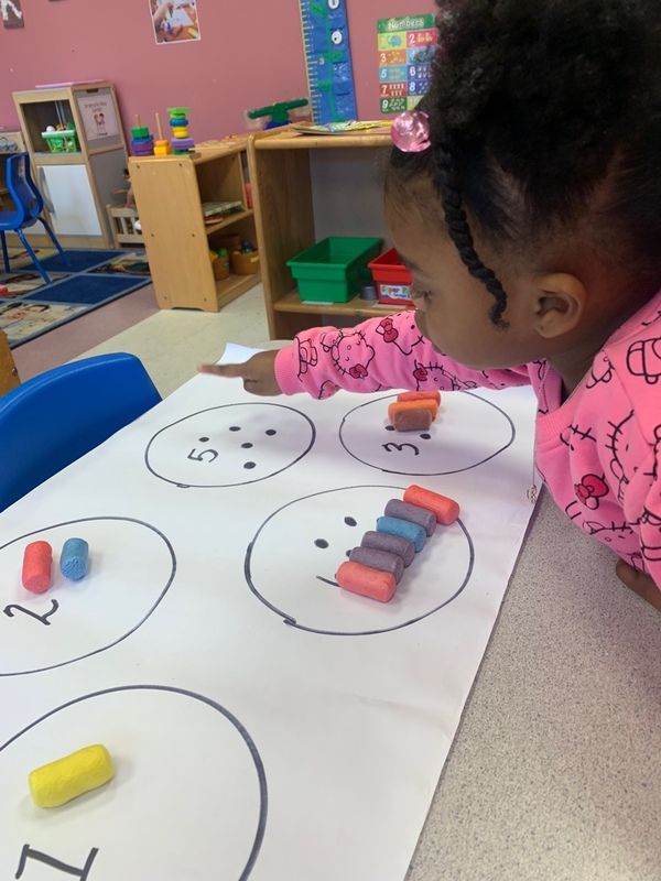 A child points to a math activity sheet on a table with circles, numbers, and colorful clay cylinders.